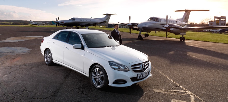 Executive Mercedes chauffeur car parked on a private airfield beside executive jets, with a uniformed chauffeur